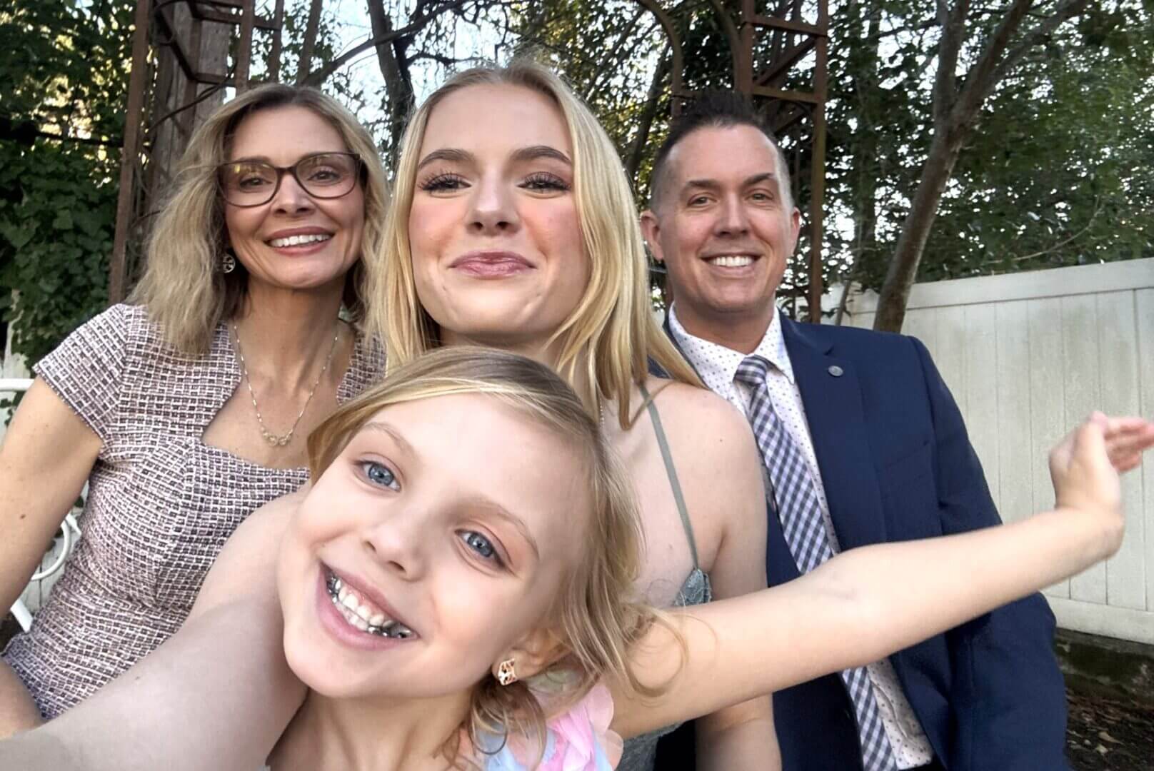 Four people, including two adults and two children, smile at the camera outdoors with trees and a white fence in the background.