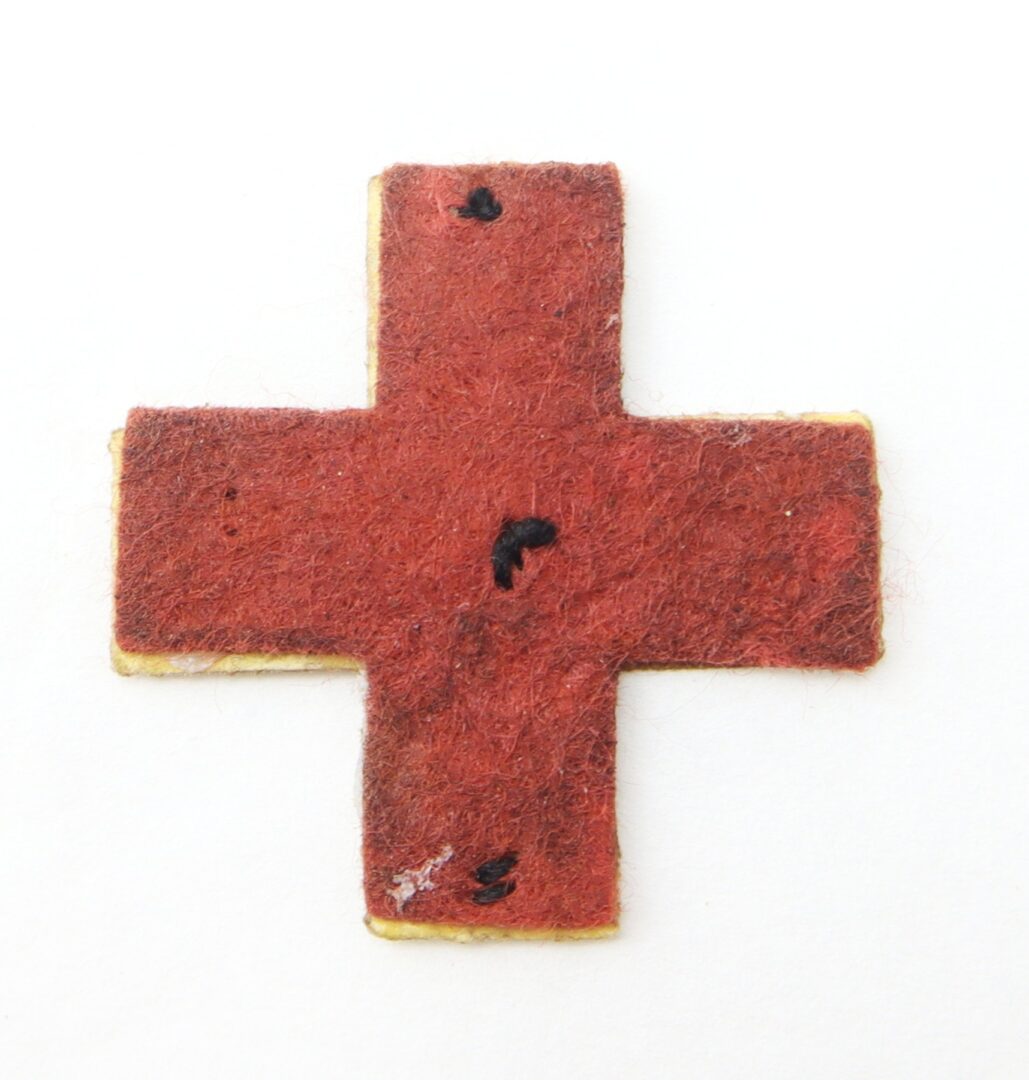 A worn Civil War Sixth Corps First Division Badge—a red wool cross with black marks and frayed edges—set against a white background.