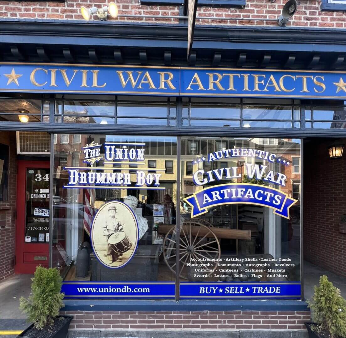 Storefront with signage reading “Civil War Artifacts,” advertising the buying, selling, and trading of items like documents, uniforms, and weapons; window displays logo and a drummer boy image.