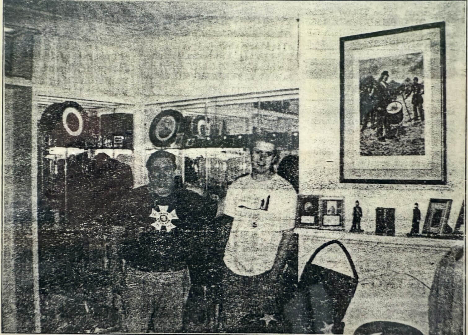 Two people stand in a room with mirrored walls, various objects on a mantel, and a framed picture on the wall above them. The image has a grainy, black-and-white appearance.