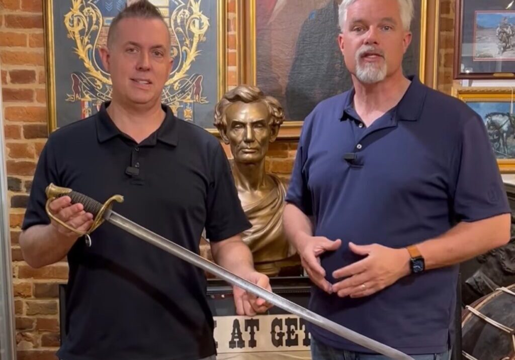 Two men stand indoors; one holds a large sword. A bust of Abraham Lincoln and various framed art pieces are visible, highlighting their collaboration with Civil War Digest.