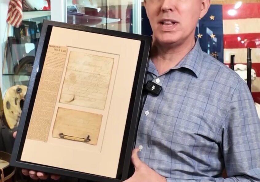 A man in a blue plaid shirt holds a framed historical document in a museum-like setting with American flags and artifacts in the background.